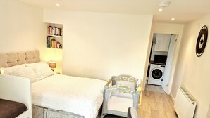 A bedroom with a bed, a small seating area, a bookshelf, a washing machine, and a radiator.