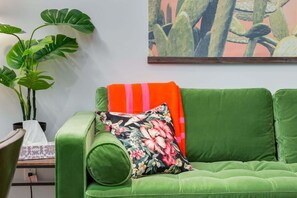 Um sofá verde com uma almofada floral e uma manta listrada, uma planta em vaso e um quadro de cactos na parede.