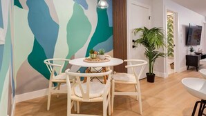 A dining area with a round table and four chairs, a large abstract mural on the wall, a potted plant, and a TV mounted on the wall. - 1BR/1BA Apartment | King Casper Bed | Sleeps 4 (Calgary)