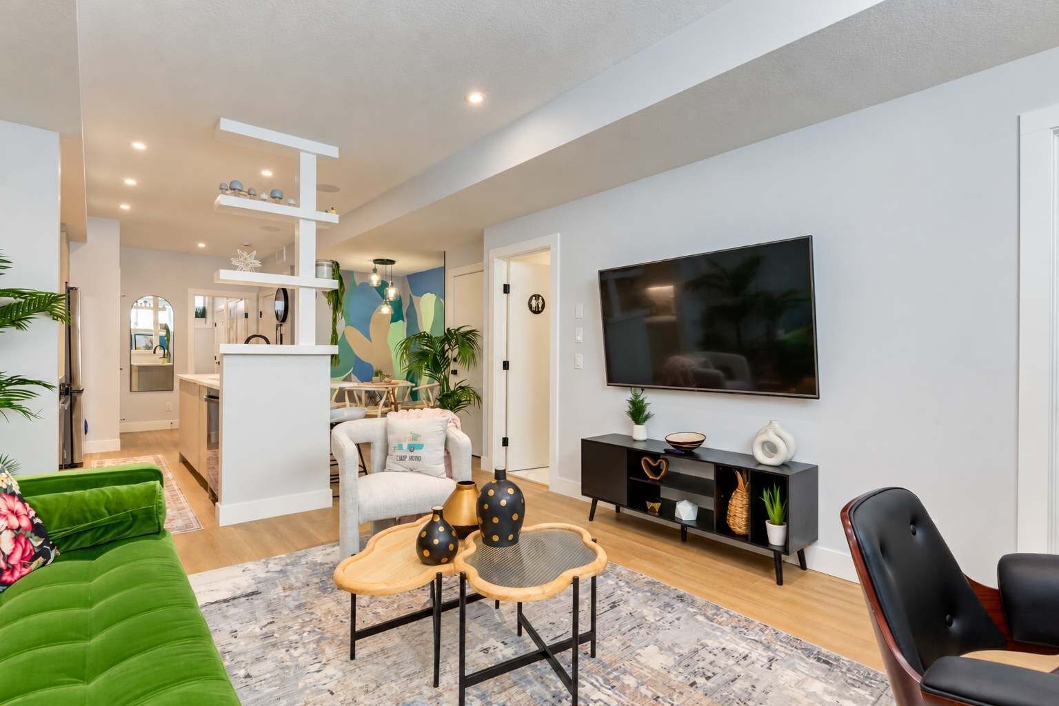 A modern living room with a flat-screen TV, a green sofa, a coffee table, and a dining area in the background.