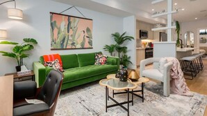 A modern living room with a green sofa, a coffee table, and a white armchair. There is a kitchen area in the background with a white staircase. - 1BR/1BA Apartment | King Casper Bed | Sleeps 4 (Calgary)