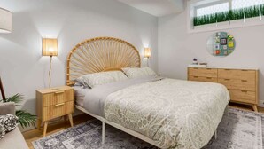 A modern bedroom with a rattan headboard, a wooden nightstand, a bedside table, a bed with patterned bedding, a round wall decoration, and a window with a view of greenery. - 1BR/1BA Apartment | King Casper Bed | Sleeps 4 (Calgary)