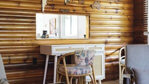Interior rústico de una cabaña con una pared de troncos de madera, un tocador blanco, una silla de mimbre con un cojín floral y un espejo que refleja una zona de cocina.