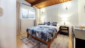 Un dormitorio con techo de madera, una cama con colcha floral, dos mesitas de noche con lámparas, una ventana con cortinas y un tapiz en la pared.