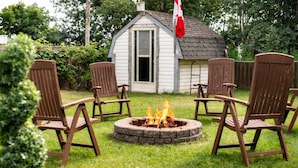 Un patio trasero con un hogar para fogata, cuatro sillas Adirondack y una pequeña casa de madera con una bandera.
