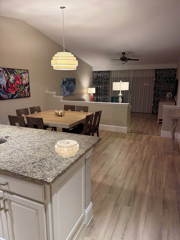 A modern kitchen with a granite countertop, wooden dining table, and chairs. There is a kitchen island with a built-in oven. The kitchen has a ceiling fan and pendant lights.