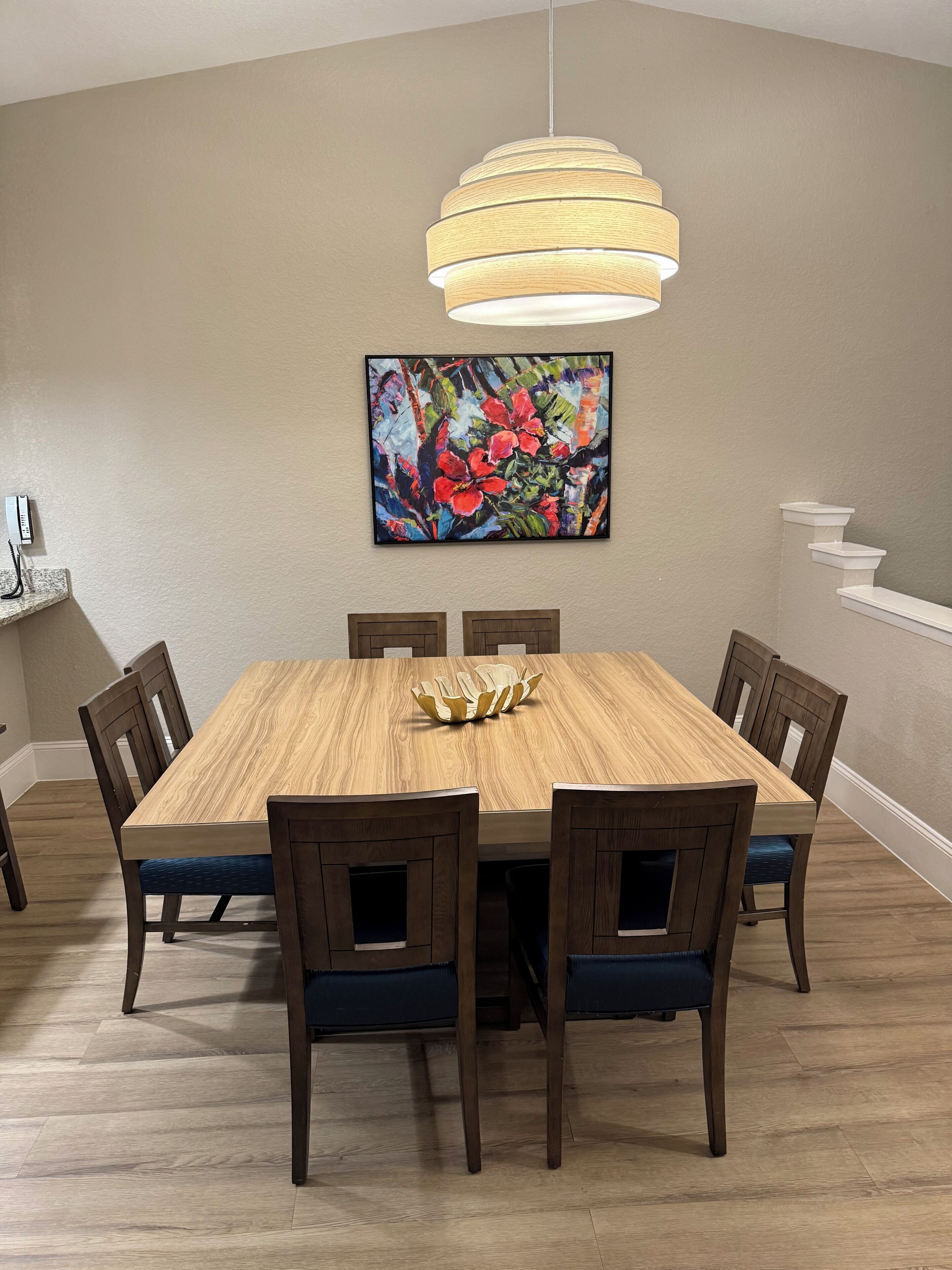 Une salle à manger avec une table en bois, six chaises, un luminaire suspendu moderne et un tableau abstrait aux couleurs vives accroché au mur.