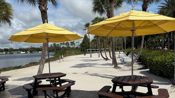 Una terraza de madera con mesas de picnic y sombrillas amarillas, con vistas a una playa con palmeras y un lago.