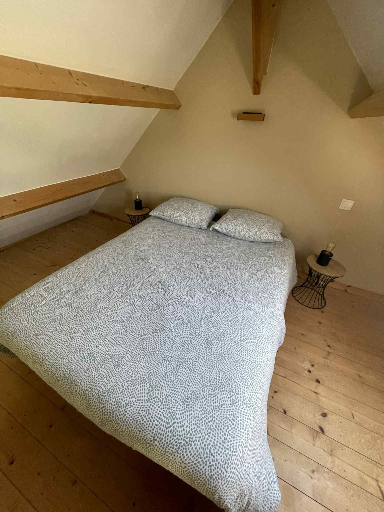 A bedroom with a sloped ceiling, wooden beams, a bed with patterned bedding, a small round table with a lamp, and a wall-mounted light switch.