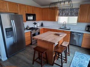 A modern kitchen with wooden cabinets, stainless steel appliances, a central island with stools, and a patterned window valance.