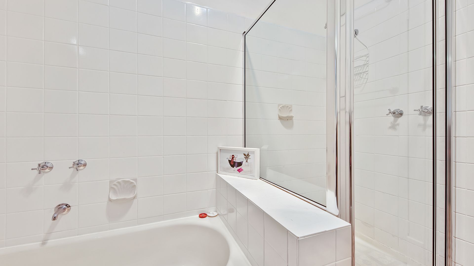 A modern bathroom with white tiles, a glass shower enclosure, a white bathtub, and a framed picture on the wall.