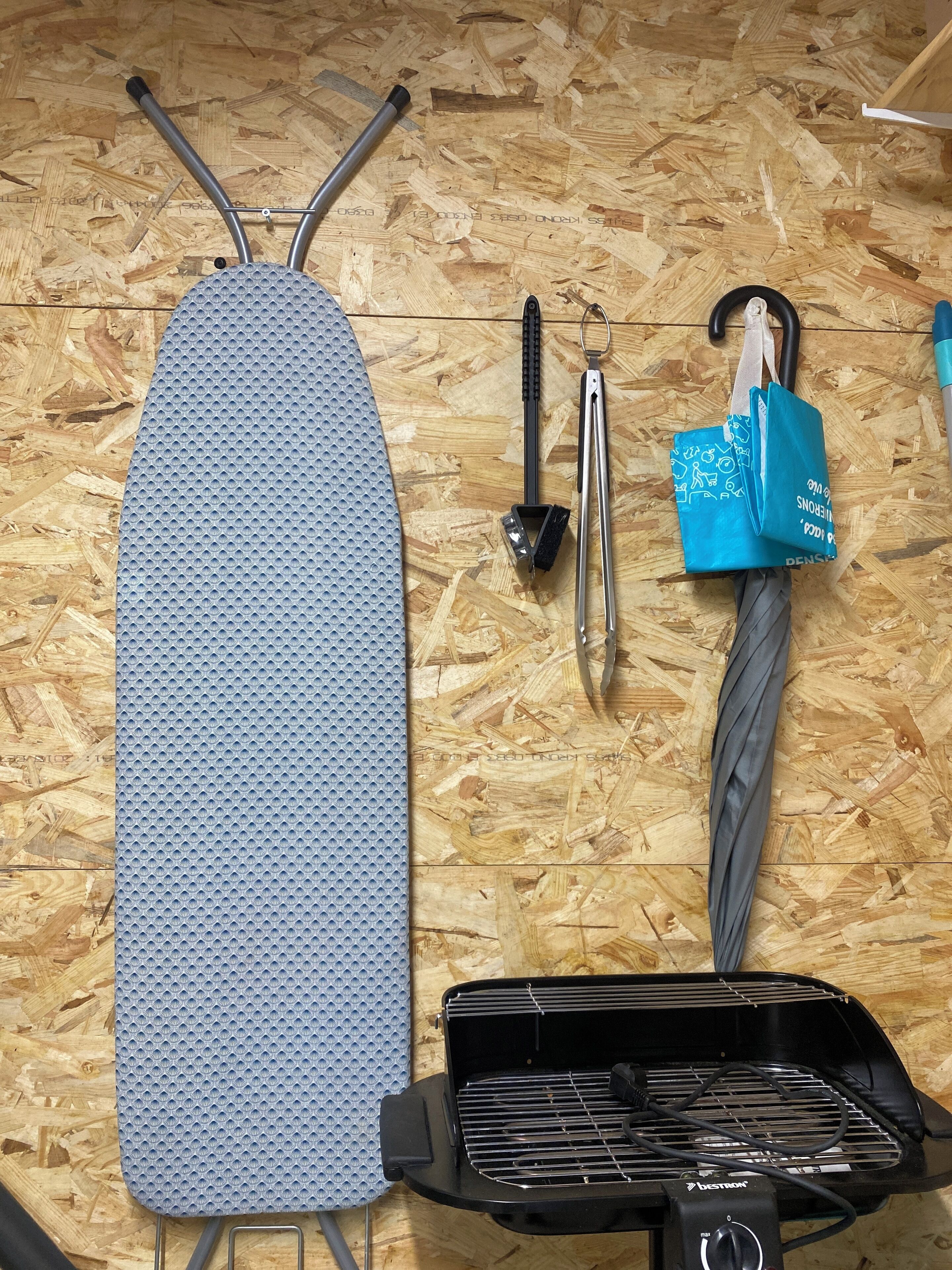 A wooden floor with a black grill, a blue ironing board, and some tools.