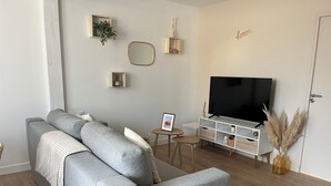 A living room with a grey sofa, a TV, a small table with a picture frame, and a wall-mounted mirror. - Private SPA & Garden - 10 min from Mont St-Michel (Pontorson)