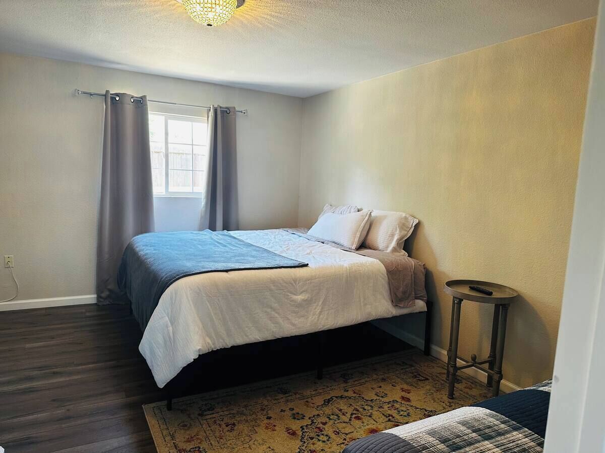 Een slaapkamer met een bed, een tafeltje, een raam met gordijnen en een kamerkleed met een patroon.