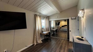 Une pièce dotée d’un plancher en bois, d’un téléviseur à écran plat fixé au mur, d’un bureau avec une chaise et d’un lit dans la pièce adjacente.