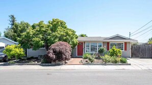 Une maison de plain-pied avec une porte rouge, un petit jardin devant avec différentes plantes et un ciel bleu dégagé.