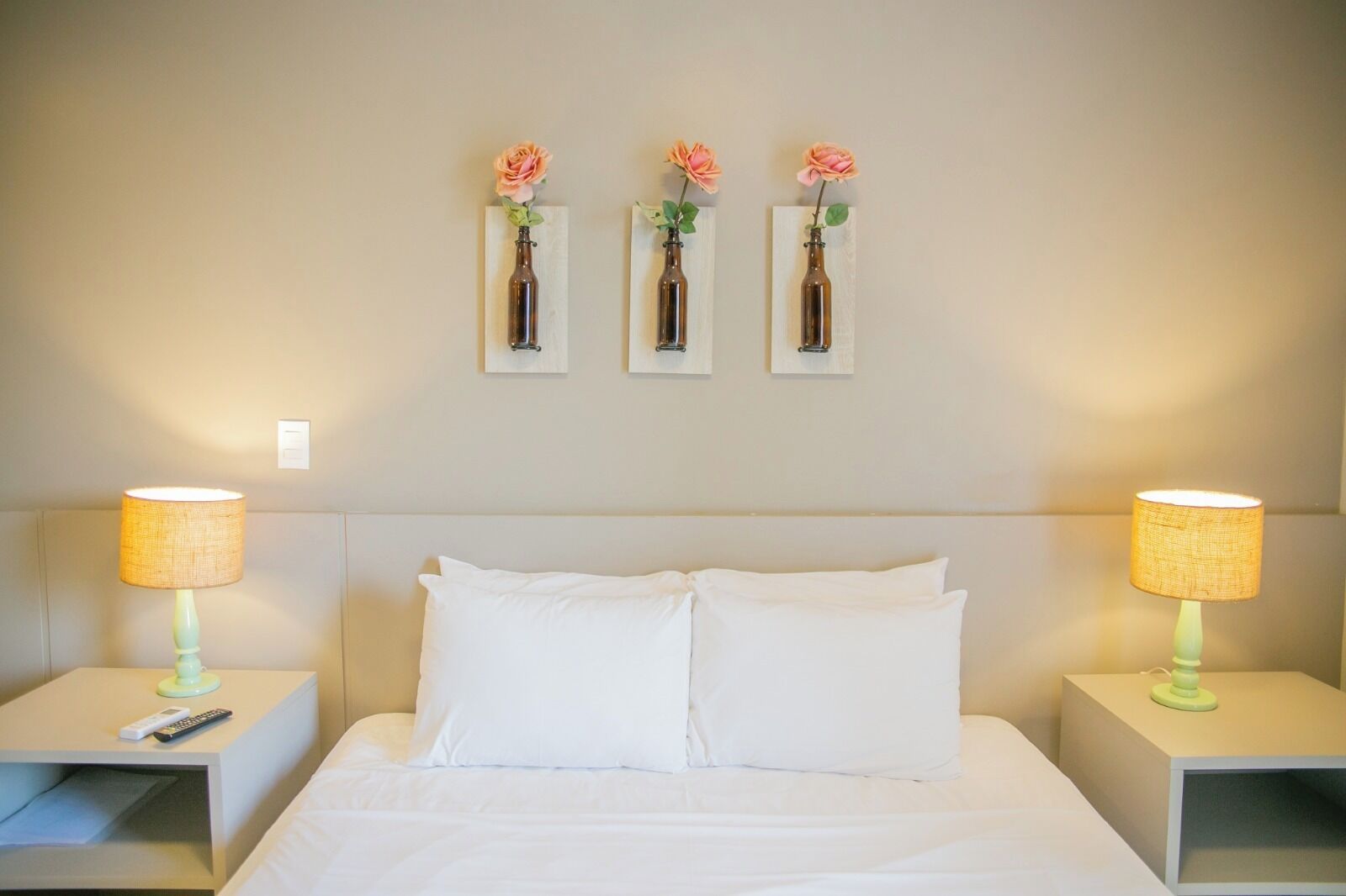 Quarto de hotel com cama, duas mesas de cabeceira, duas luminárias e três vasos de flores fixados na parede.