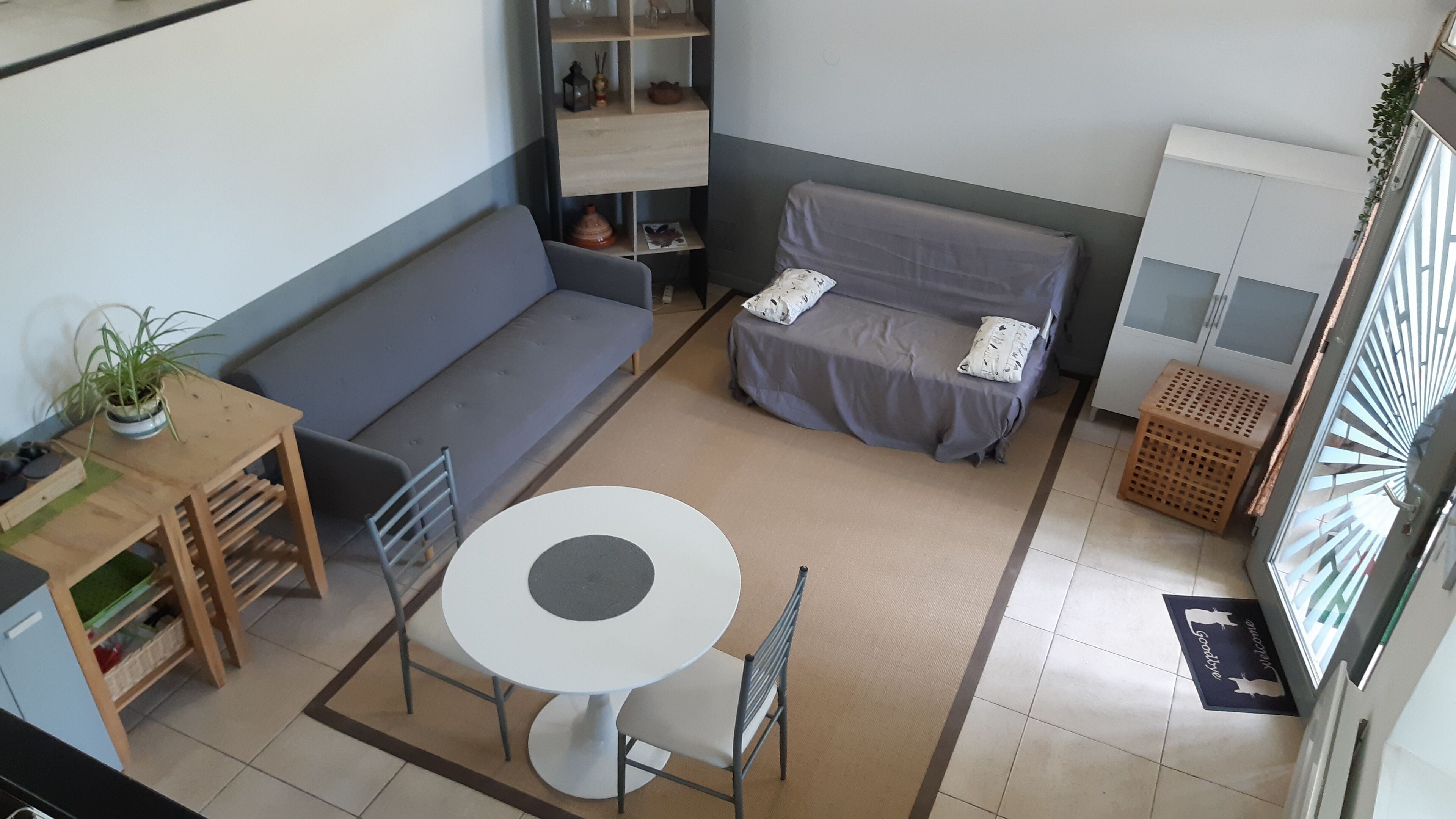 Un espace de vie compact comprenant un canapé, une petite table ronde, une chaise, une table d’appoint en bois avec une plante, un meuble et un panier à linge.