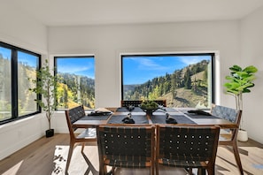 A dining area with a table set for six, large windows offering a view of a forested mountain range.