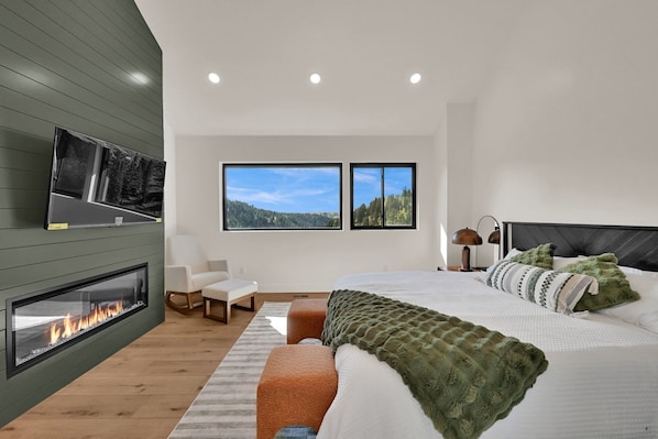 A modern bedroom with a fireplace, a bed with green bedding, a large window with a view of trees, and a white chair.