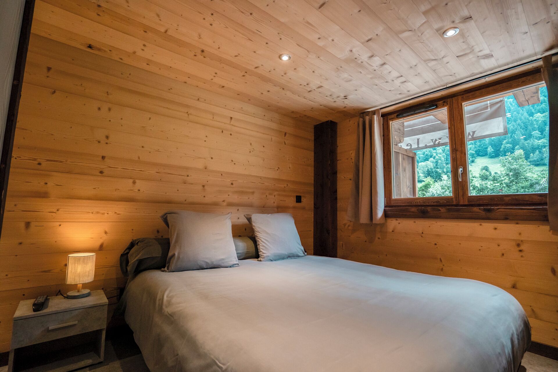 A wooden cabin bedroom with a bed, pillows, a nightstand with a lamp, and a window with a view of greenery.