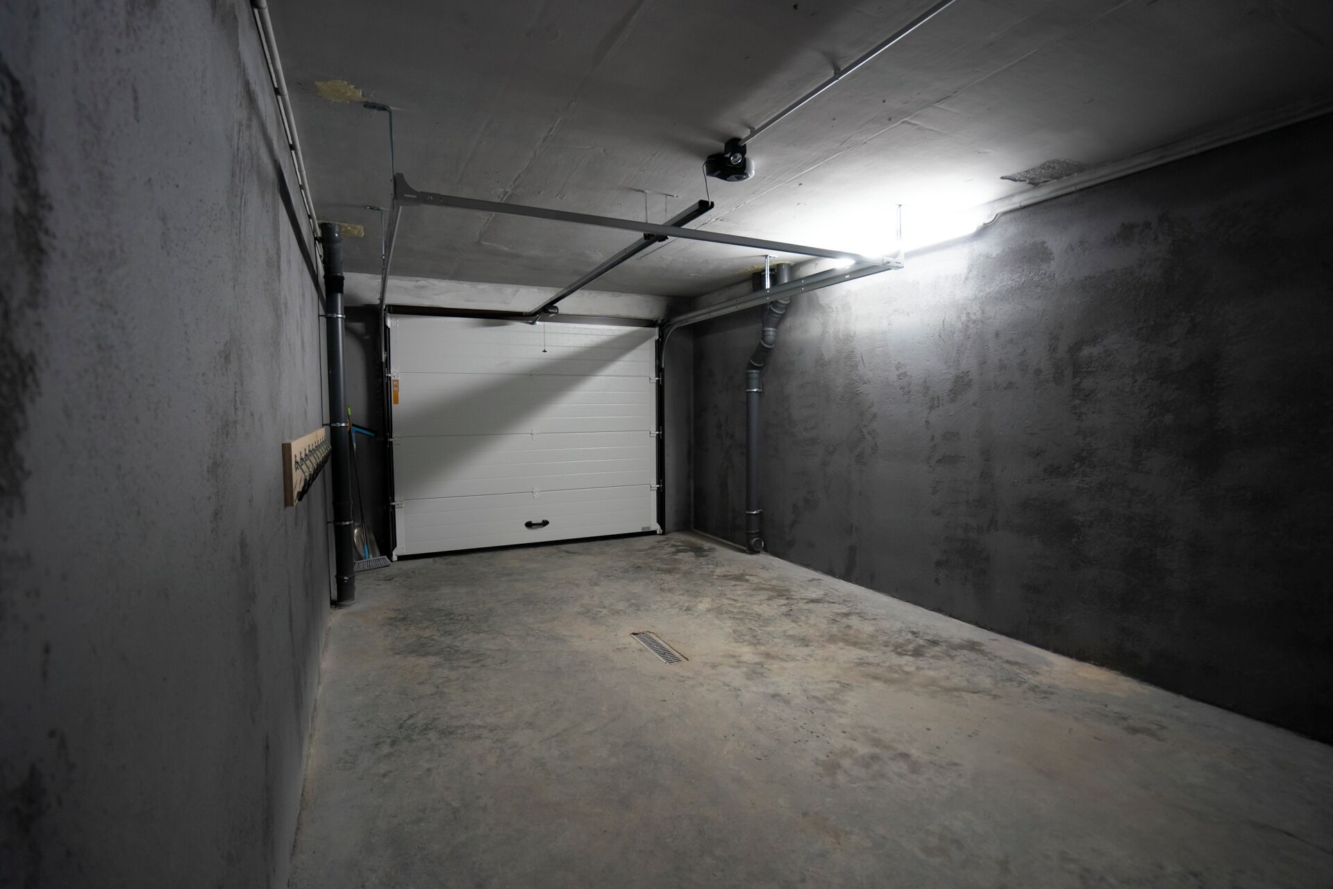 Et svagt oplyst garage med en lukket hvid dør, betonvægge og synlige rør.