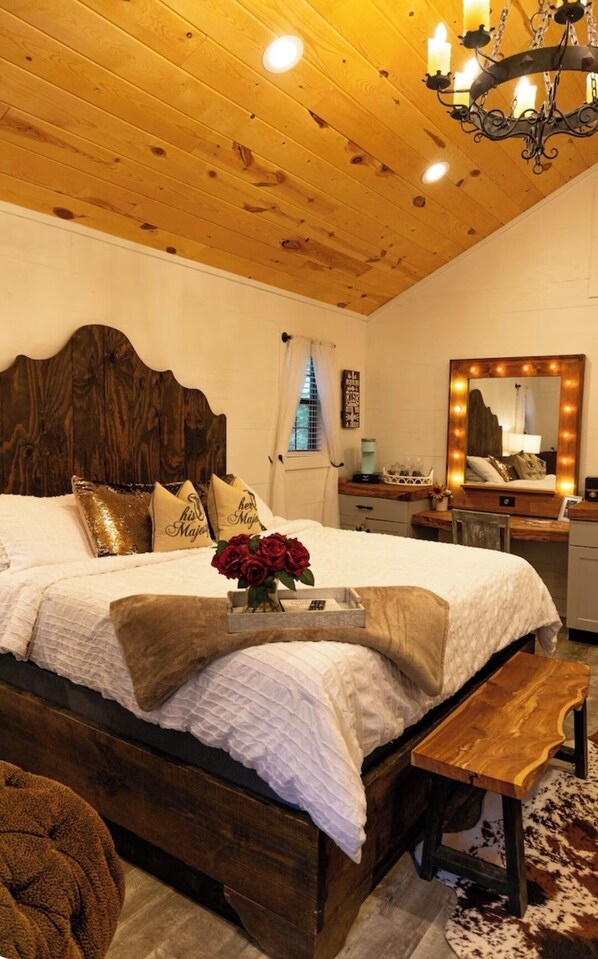 Une chambre confortable avec un plafond en bois, un grand lit avec une literie blanche, un banc en bois, une coiffeuse avec un miroir et un lustre.