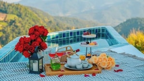 A dining setup with a table, a vase of red roses, a teapot, and a tray with food items, set against a mountainous backdrop.