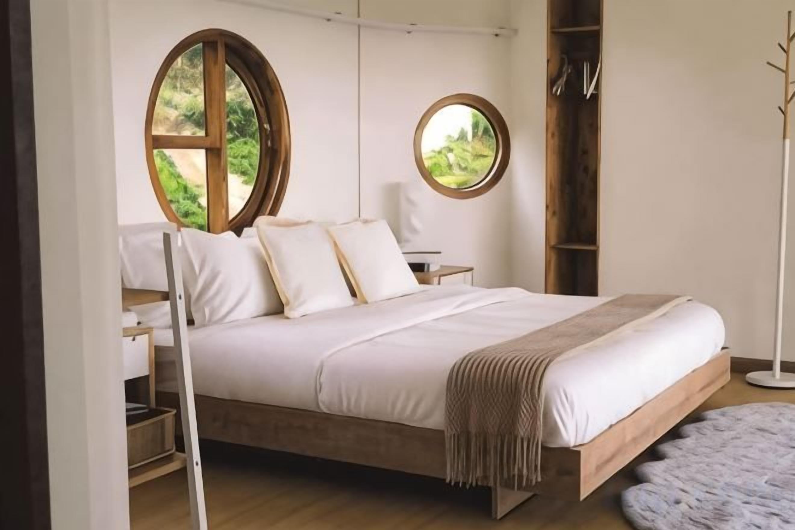 A bedroom with a wooden bed, two round windows, a bedside table, and a floor lamp.