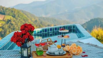 Ein Tisch ist mit einem Strauß roter Rosen, einem Teekessel, Tassen und einer Auswahl an Gebäck gedeckt, mit einer Berglandschaft im Hintergrund.