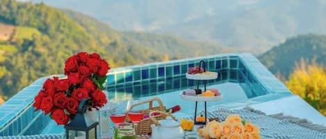 A table set with a bouquet of red roses, a teapot, cups, and a tray of pastries, with a mountainous backdrop.