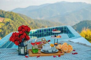 Ein Tisch ist mit einem Strauß roter Rosen, einem Teekessel, Tassen und einer Auswahl an Gebäck gedeckt, mit einer Berglandschaft im Hintergrund.