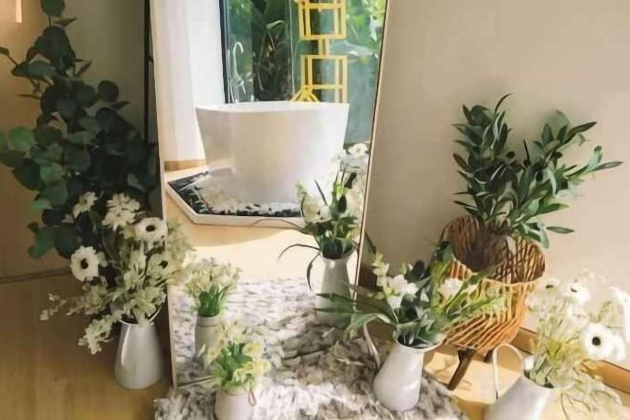 Et værelse med et stort spejl, der viser et badekar og et vindue med en gul stige. Der er flere krukkeplanter og blomster placeret på gulvet.