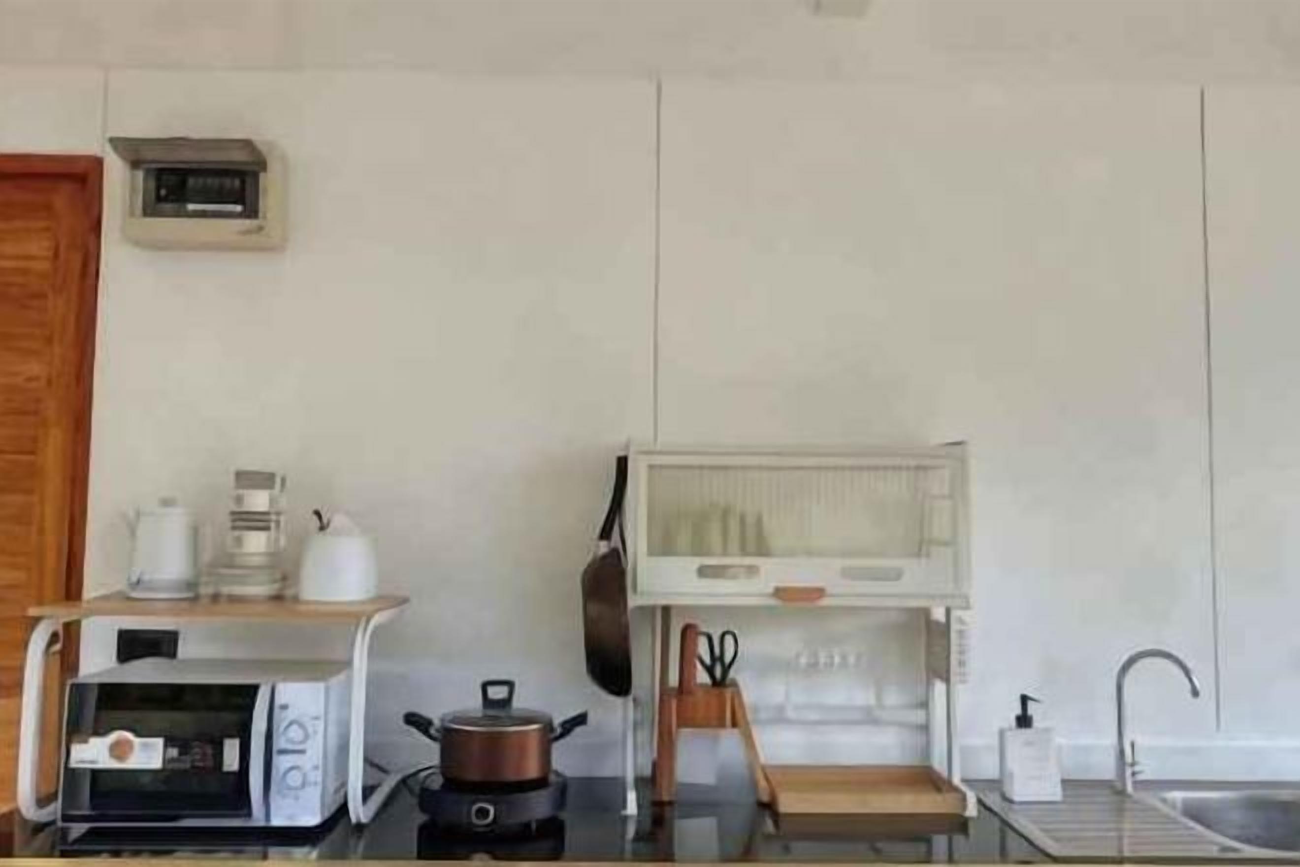 A compact kitchen with a microwave, kettle, and kettle on a shelf, a pot on the counter, a sink, and a clock on the wall.