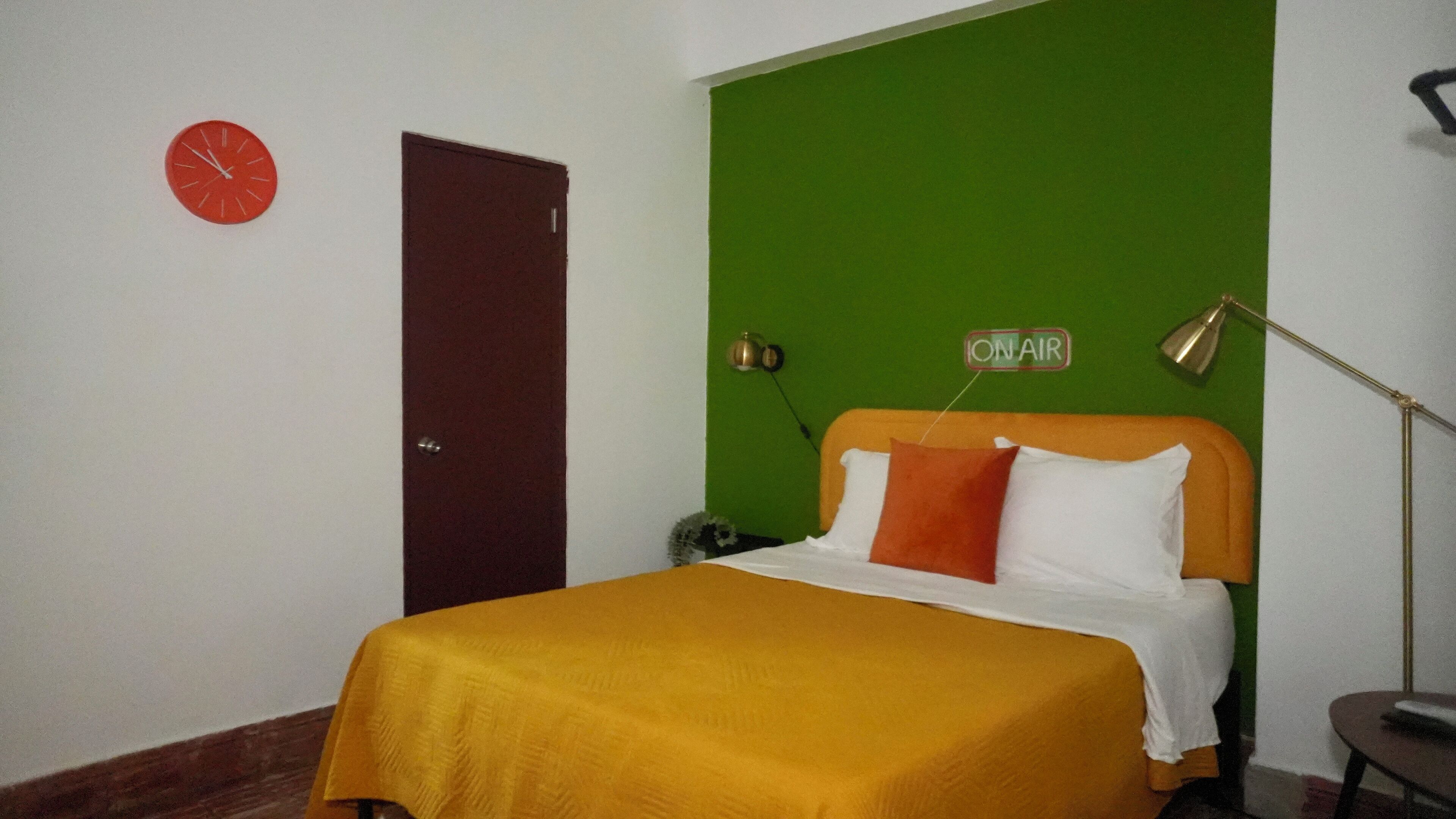 A room with a bed, a red clock, a green wall, a door, and a lamp.