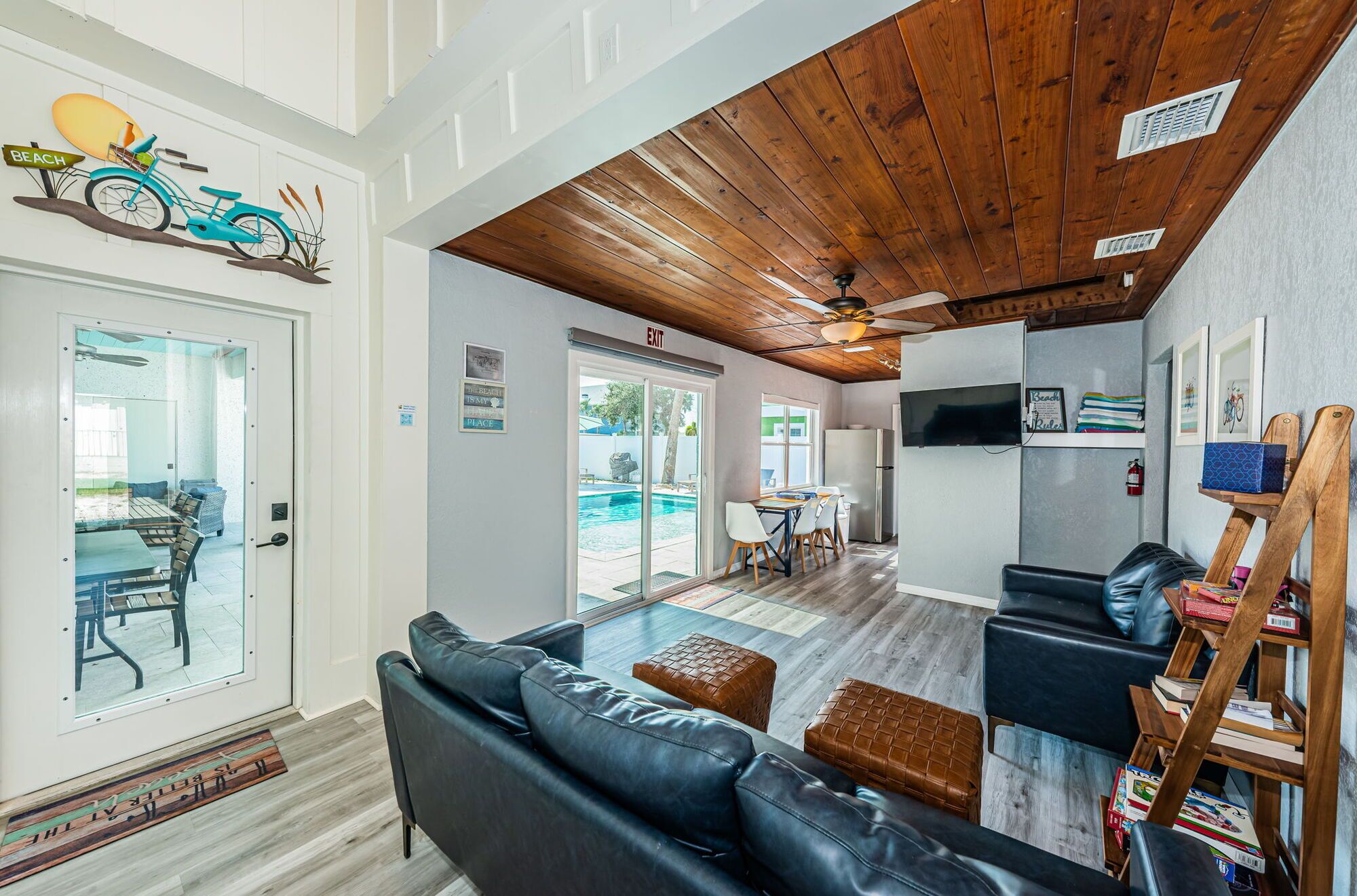 A modern living room with a wooden ceiling, a pool view, and a beach-themed sign.