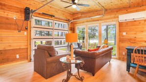 L’intérieur d’un chalet en bois, avec deux canapés, une table en verre, un ventilateur de plafond et de grandes fenêtres.