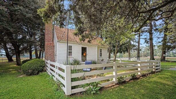 A white wooden house with a brick chimney, surrounded by a white picket fence and lush green grass.