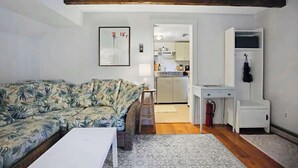 A living room with a sofa, a small table, and a white cabinet. There is a kitchen visible through an open doorway.