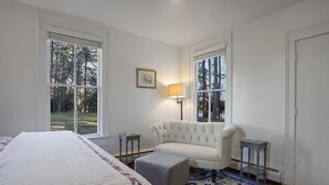 Een slaapkamer met een groot bed, een gestoffeerde fauteuil, een klein voetenbankje, twee nachtkastjes en een lamp.