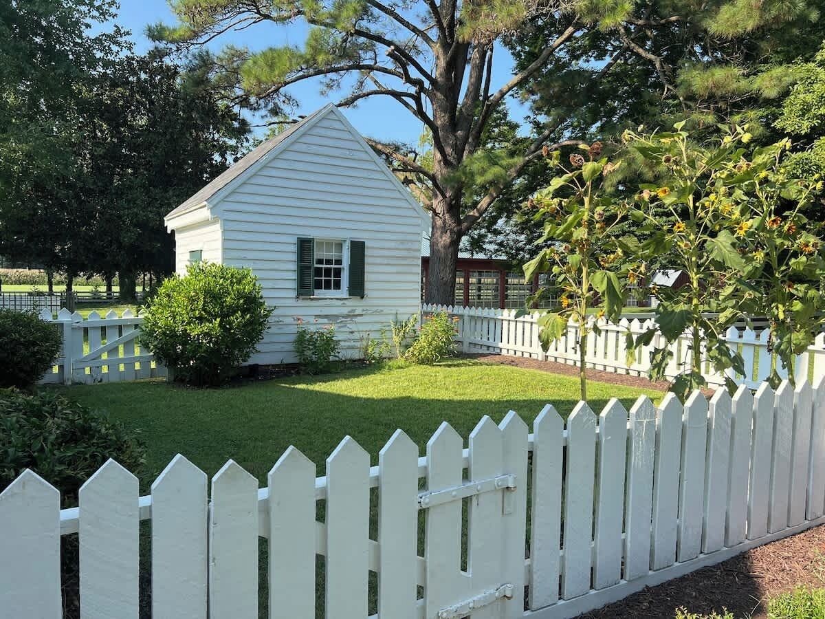 A white house with a white picket fence, surrounded by green grass and trees.