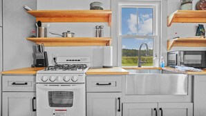 A modern kitchen with a gas stove, oven, stainless steel cabinets, wooden shelves, and a window with a view of greenery.