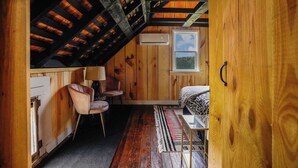 A cozy attic bedroom with wooden walls, a sloped ceiling, a bed, a chair, a small table, and a window with a view.
