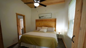 Une chambre à coucher avec une tête de lit en bois, un lit recouvert d’une couvre-lit verte et blanche, un ventilateur de plafond et une fenêtre avec des stores.