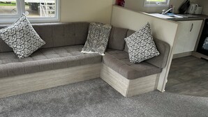 A compact living space with a grey sectional sofa, patterned cushions, a small kitchen area with a microwave, and a window with a view of a house. - 3 bed mobile home - Holiday Park (Northampton)