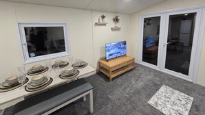 A dining area with a table set for four, a television on a stand, a bench, and a window with a view of the outside. - 3 bed mobile home - Holiday Park (Northampton)