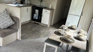 A compact kitchenette with a microwave, stove, and white cabinets. A dining table with plates and glasses is set up. - 3 bed mobile home - Holiday Park (Northampton)