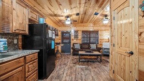 A cabin interior with wooden walls and floors, a kitchen with granite countertops, a refrigerator, and a dining area with a table and chairs. - Rustic Retreat W/ Hot Tub • Firepit - BBQ (Broken Bow)
