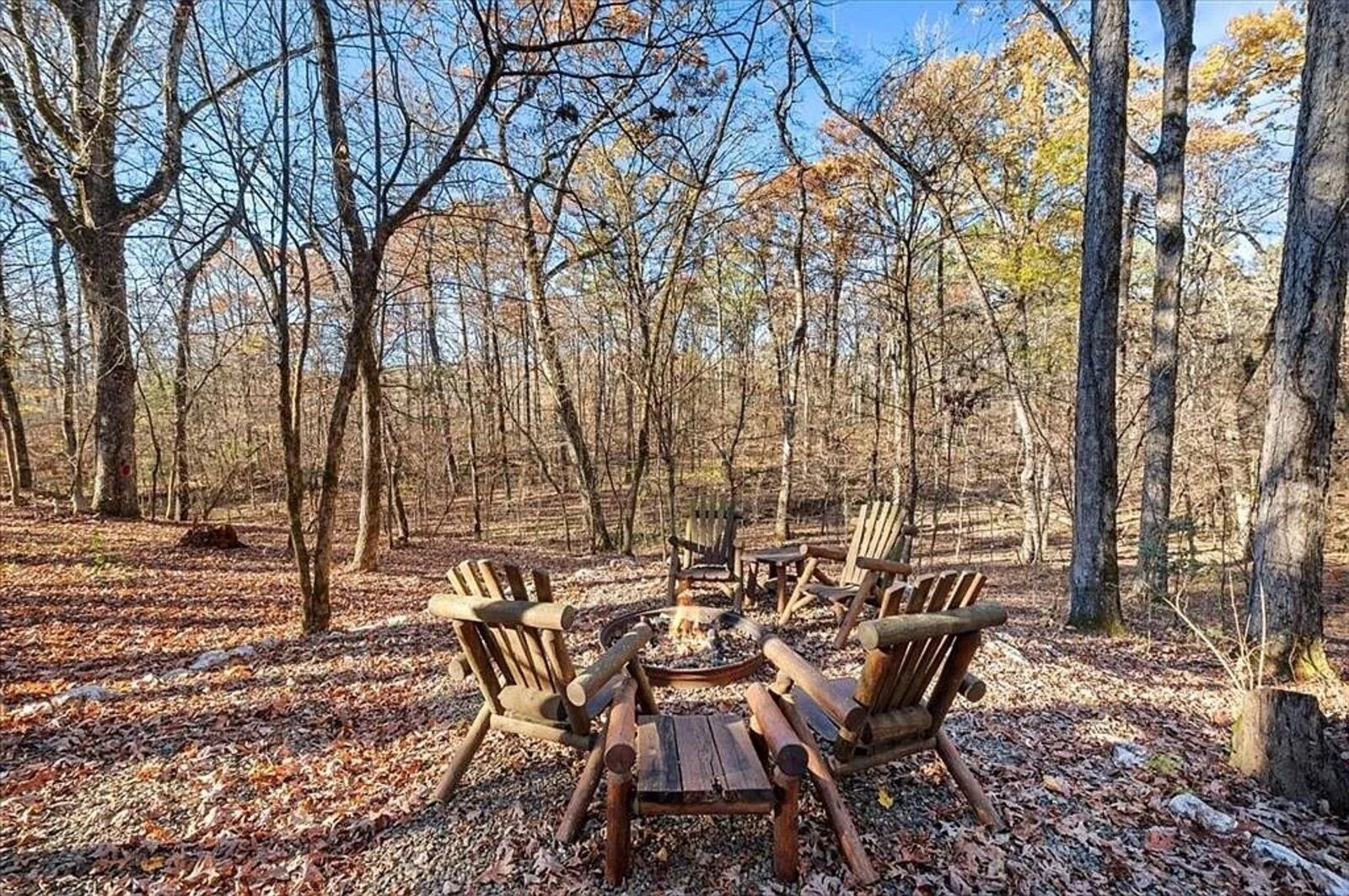 A forest clearing with a wooden table and four Adirondack chairs.