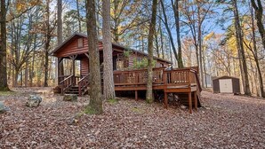 A cabin with a porch, a deck, and a shed in a wooded area. - Rustic Retreat W/ Hot Tub • Firepit - BBQ (Broken Bow)